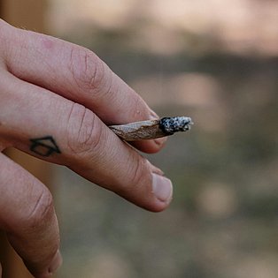 Abgebildet ist die Hand eines jungen Menschen mit einem Joint zwischen den Fingern